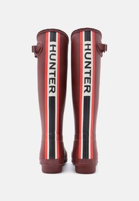 Raudoni guminių pirštinių batai su baltais ir juodais vertikaliais dryžiais, ant kurių bold rašmenimis parašyta "HUNTER", ir šone esantis dirželis reguliavimui.