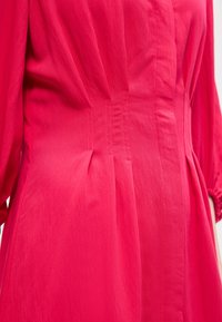 Εγγύς πλάνο φωτεινού ροζ υφάσματος με λεπτομερή ραφές και πιέτες σε ένδυμα, που αποκαλύπτει την υφή και τις γραμμές της ραφής.