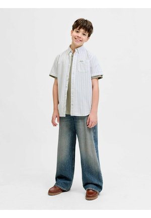 Un garçon souriant debout portant une chemise légère à manches courtes rayée par-dessus un t-shirt vert, un jean délavé à jambes larges et des chaussures marron sur un fond blanc.