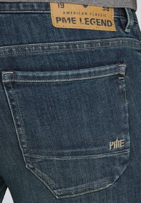 Donkerblauwe denim jeans met een getextureerde afwerking. Voorzien van een bruine leren label en geborduurde branding op de achterzak.