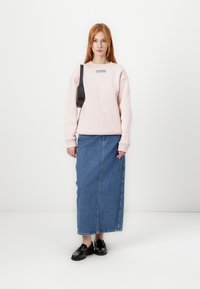 Felpa rosa con un piccolo logo nero, abbinata a una lunga gonna di jeans blu. La modella indossa mocassini neri. Sfondo semplice.