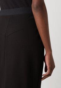 Robe noire avec une texture lisse et une large taille élastique ; présente des poches en seam incurvées à l'arrière pour un look sur mesure.