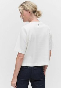 Valkoinen lyhythihainen cropped-toppi, jossa on pyöreä pääntie ja pieni musta nappi takana. Pehmeä kangastuntuma.