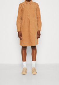 Beige Cord-Shirtkleid mit Kragen, Knopfverschluss und einer Brusttasche. Lange Ärmel, knielang, kombiniert mit weißen Socken und beigen Schuhen.