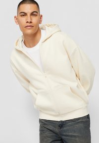 Haut à zip crème avec une poche kangourou, tissu léger, coupe décontractée et capuche à cordon, associé à un jean foncé.