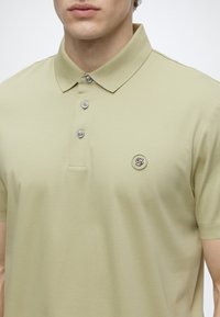 Homme portant un polo vert olive clair avec trois boutons et un petit logo rond sur la poitrine.