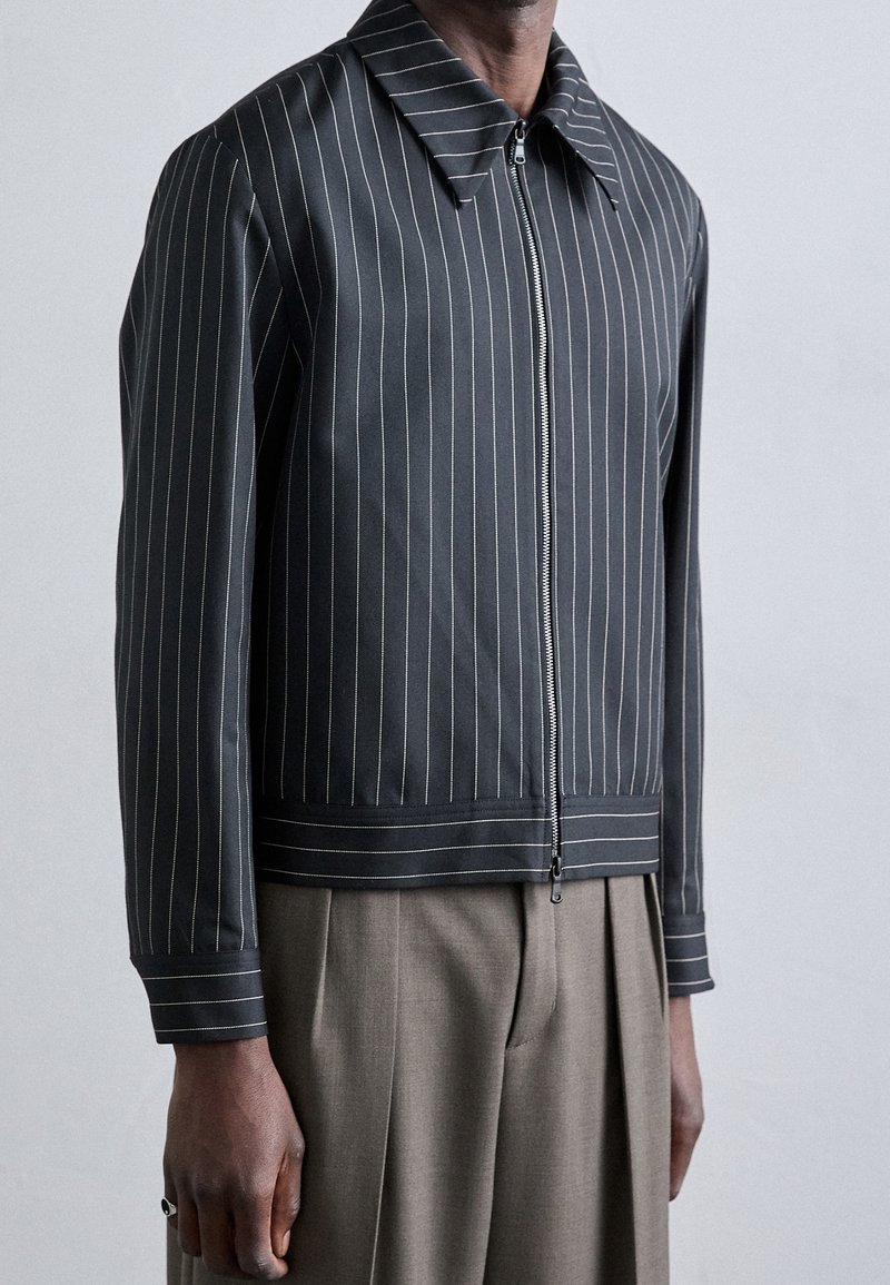 Homme portant une veste noire à fines rayures zippée avec un large col et un pantalon plissé beige sur un fond uni.