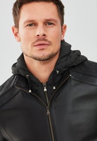 Veste en cuir noir au design ajusté, dotée d'une fermeture éclair et d'accents côtelés sur les épaules. Portée par-dessus un sweat à capuche gris foncé.