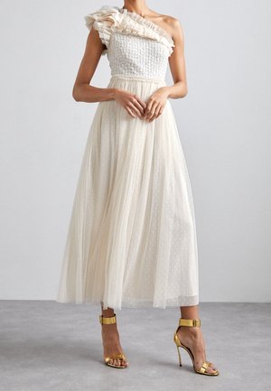 Vrouw draagt een crèmekleurige off-shoulder jurk met ruches en een tule rok, gecombineerd met gouden sandalen met hoge hakken, staand op een grijze vloer.