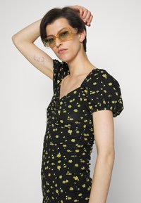 Vestido preto floral com mangas curtas em balão, com flores amarelas. Óculos de sol transparentes amarelos complementam o visual. Textura do tecido suave.