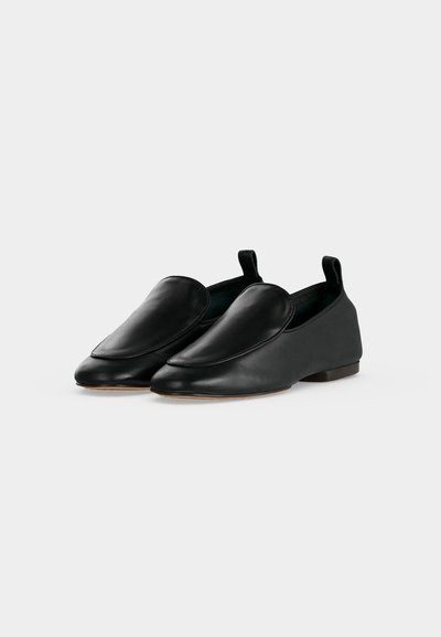 Ζευγάρι μαύρα δερμάτινα παπούτσια τύπου slip-on με επίπεδη σόλα, μυτερές μύτες και πίσω θηλιές, εμφανιζόμενα μπροστά σε απλό ανοιχτό γκρι φόντο.