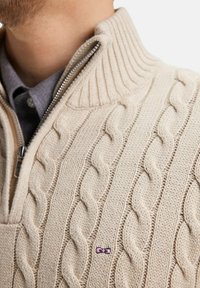 Pull en tricot beige avec un col large côtelé, doté d'une demi-fermeture éclair et d'un petit logo en forme de nœud violet sur la poitrine.