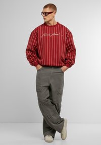 Červený pruhovaný sveter s bielymi pruhmi; šedé cargo nohavice; béžové tenisky; model stojí s rukami v vreckách; elegantné slnečné okuliare dopĺňajú vzhľad.