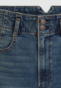 Jeans de ganga azul de cintura alta com um cós em forma de V, rebites de cobre e pespontados laranja em contraste, com uma textura desbotada.