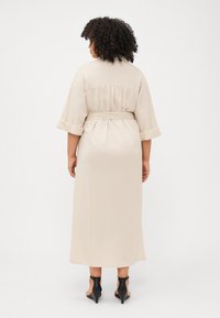 A person with curly hair wears a beige, belted maxi dress, facing away. Black heeled shoes are visible at the feet. Neutral background.