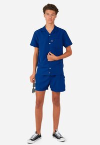 Blauw overhemd met korte mouwen en bijpassende shorts van gladde stof, met een borstzak en witte knopen. Gemaakt om te dragen met zwarte sneakers.