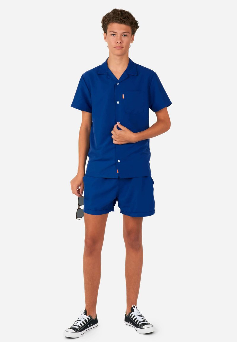 Blauw overhemd met korte mouwen en bijpassende shorts van gladde stof, met een borstzak en witte knopen. Gemaakt om te dragen met zwarte sneakers.