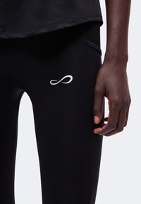 Gros plan de la main droite d'une personne à côté d'un legging de sport noir avec un petit logo infini blanc sur la cuisse supérieure.