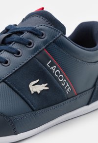 Baskets bleu marine en textile et daim, avec une semelle blanche, des accents rouges et un logo crocodile argenté sur le côté.