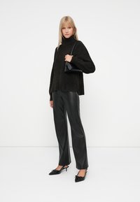 Überdimensionierter schwarzer Pullover mit strukturiertem Strick, kombiniert mit hochgeschnittenen schwarzen Lederhosen und schwarzen spitzen Absatzschuhen. Akzentuiert mit einer kleinen Tasche.