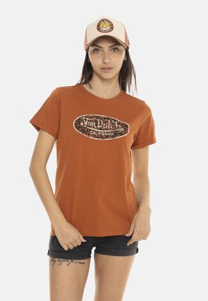Jeune femme portant un t-shirt orange Von Dutch California, un short noir et une casquette camionneur Von Dutch rose et beige, debout sur fond blanc.