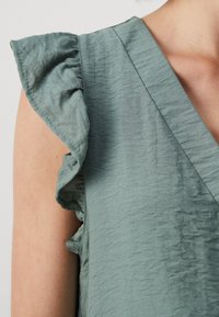 Haut vert menthe avec manches volantées. Présente un décolleté en V et un tissu texturé, mettant en valeur de douces plis. Vue rapprochée de la zone de l'épaule.