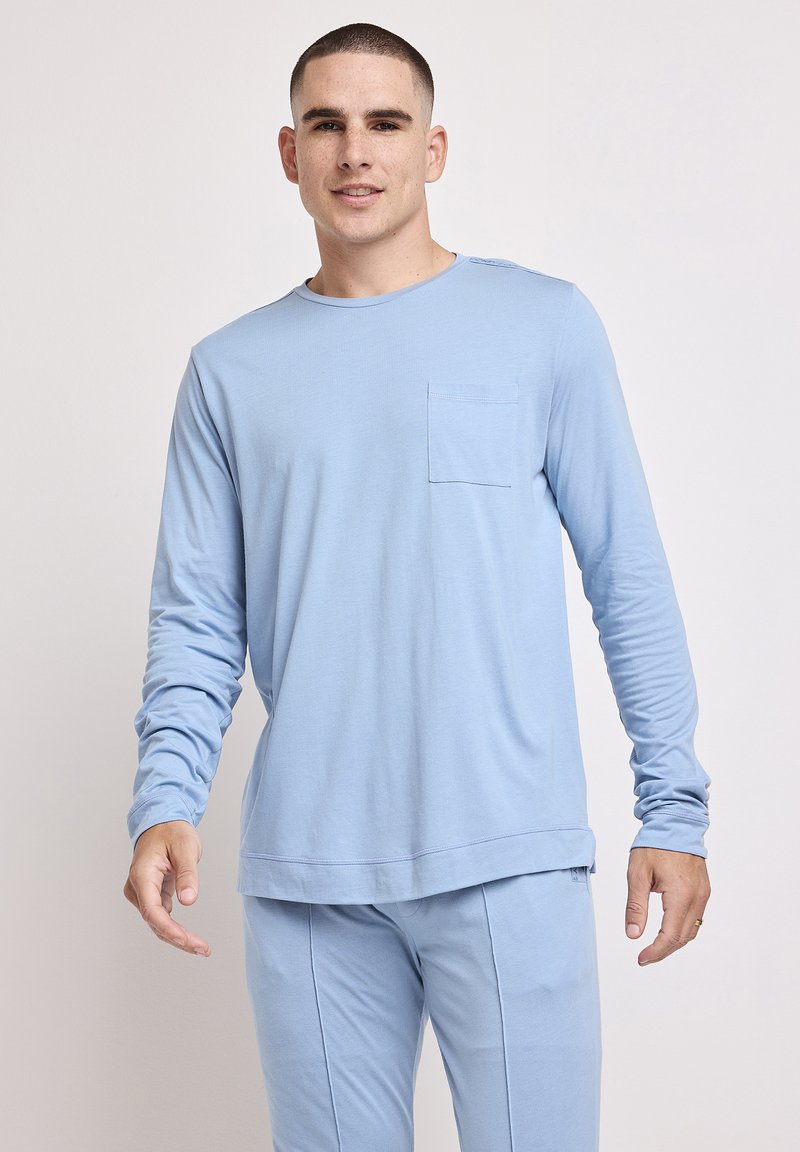 Langærmet lyseblå skjorte lavet af blødt stof, med en rund halsudskæring og en brystlomme, båret med matchende lyseblå bukser.