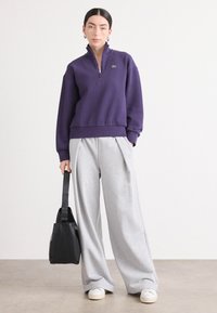 Paarse ritsvest met hoge kraag, gecombineerd met losse grijze broek en witte sneakers, met een zwarte handtas in de hand.