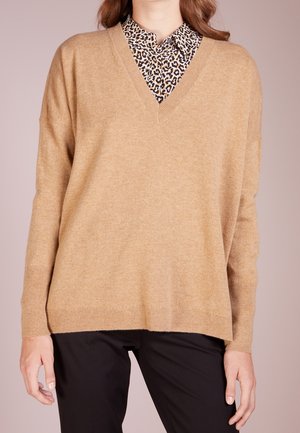Beige v-ringad tröja gjord av mjukt stickat tyg, med sänka axlar och ribbade muddar, lagd över en leopardmönstrad skjorta med krage.