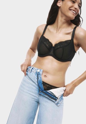 Vrouw die glimlacht terwijl ze lichtblauwe jeans over zwarte kanten onderbroek en bijpassende bh trekt, tegen een witte achtergrond.