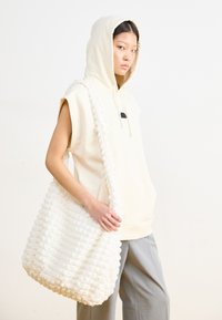 Weiße strukturierte Handtasche aus weichem Stoff, mit einem geflochtenen Tragegurt, getragen von einem Modell in einem hellbeigen Hoodie und grauen Hosen.