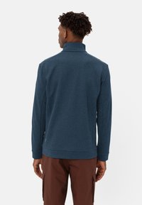 Giacca a pullover blu navy con colletto alto, tessuto a trama e maniche lunghe. Pantaloni cargo marroni visibili, con un sottile dettaglio del logo verde.