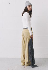 Valkoinen pitkähiha, vaaleanruskeat corduroy-laskoslahkeet, harmaa neulepipo, pitää tummanharmaata kuviollista villapaitaa. Yksinkertainen tausta.