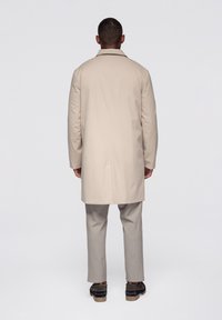 Gabardina beige con cuello clásico, mangas largas y corte recto, combinada con pantalones gris claro y zapatos oscuros.