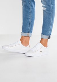 White synthetic sneakers with a smooth texture, featuring a tan accent on the side and laces. Worn with light blue cuffed jeans.