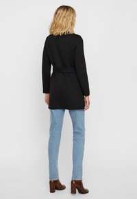 Black belted coat with long sleeves, straight silhouette, knee-length design, paired with light blue jeans and brown heeled boots.