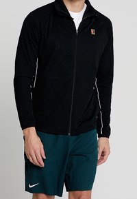 Uomo che indossa una giacca nera con zip e un piccolo logo sul petto, insieme a pantaloni corti Nike verde scuro, su uno sfondo semplice.