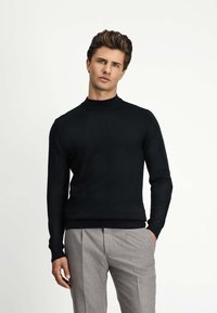 Czarny dopasowany sweter z wysokim kołnierzem i długimi rękawami, wykonany z gładkiego materiału. W zestawieniu z jasnoszarymi, plisowanymi spodniami.