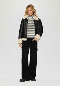 Veste en cuir noir avec un col en shearling crème, associée à un pull col roulé noir et blanc à rayures et un pantalon cargo noir à jambes larges. Bottes noires.