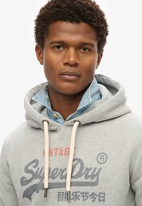 Grå hoodie med snore, der har rød og marineblå branding. Båret over en lys blå, denimskjorte med knapper og en tekstureret krave.
