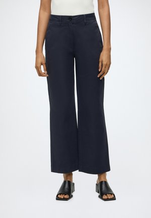 Persoon draagt een donkere, wijde broek, wit mouwloos topje en zwarte sandalen met hak en staat tegen een effen lichte achtergrond.
