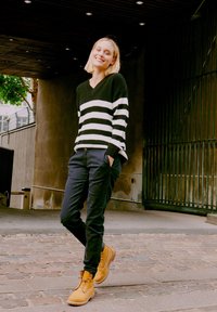 Fekete-fehér csíkos pulóver, laza fekete nadrág és bézs fűzős csizma. Egy texturált falnak támaszkodva, modern háttérrel.