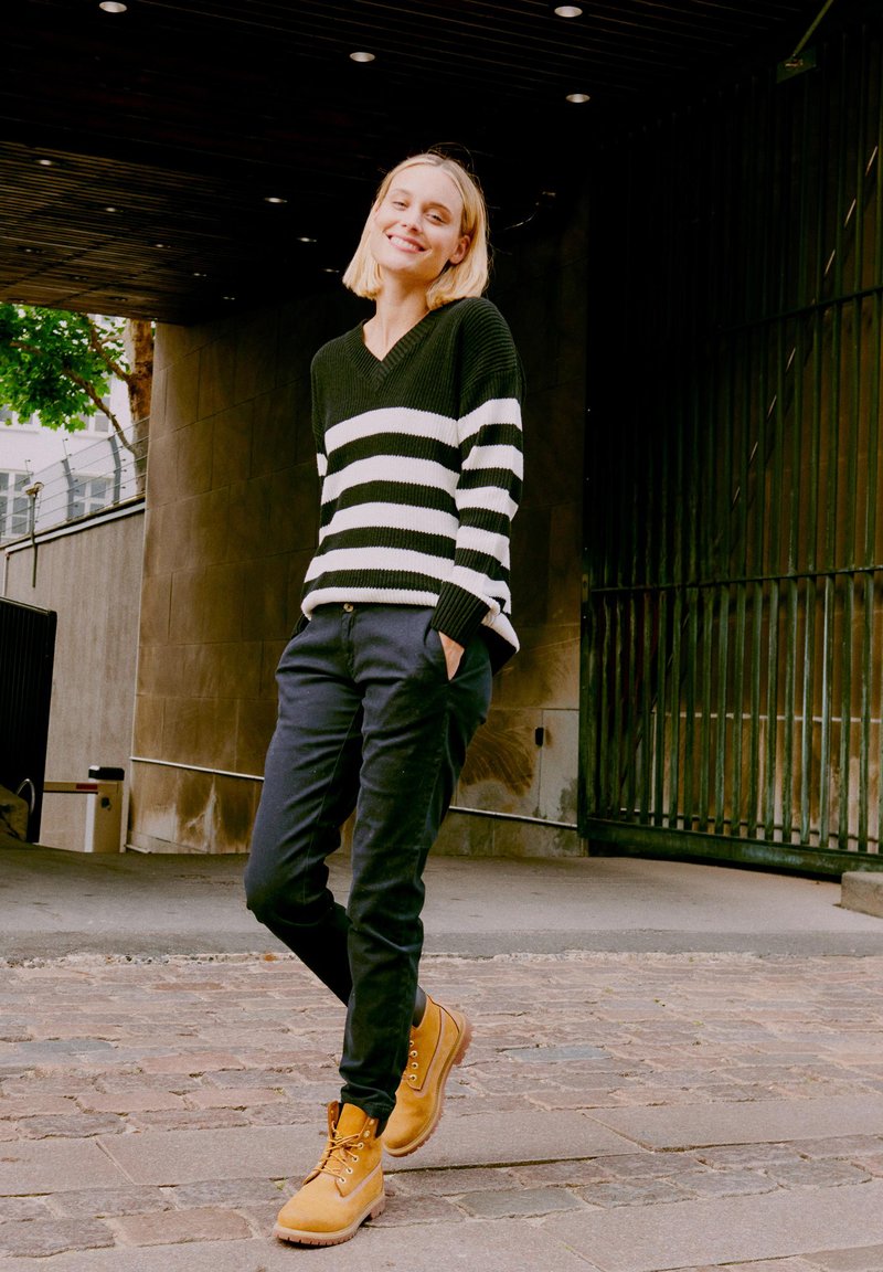 Fekete-fehér csíkos pulóver, laza fekete nadrág és bézs fűzős csizma. Egy texturált falnak támaszkodva, modern háttérrel.