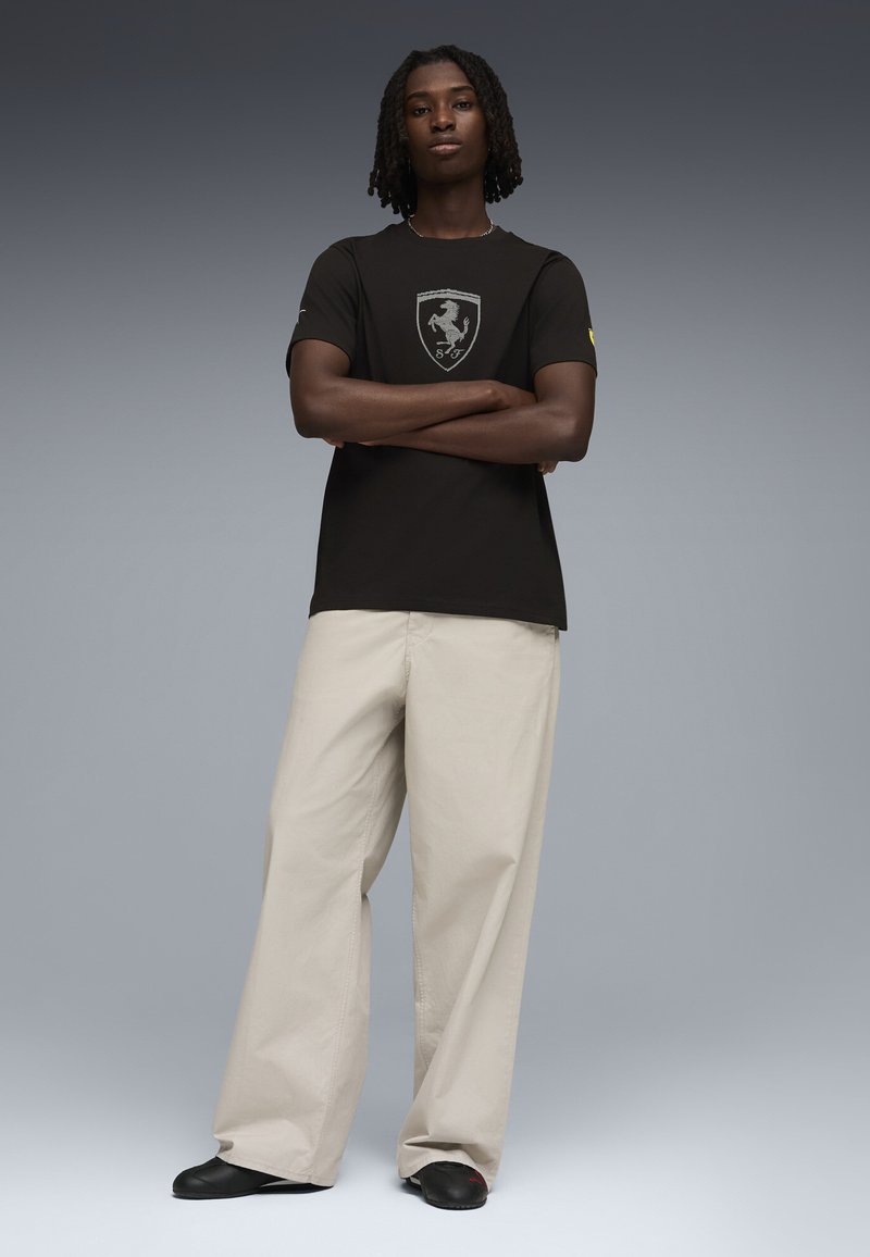 Young person with crossed arms wearing black Ferrari logo t-shirt, beige wide-leg trousers, and black shoes standing on gray background.