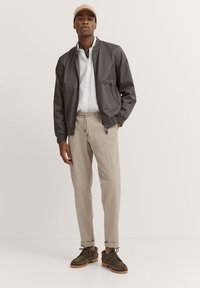 Graue Bomberjacke mit zwei Fronttaschen, weißes Hemd darunter, beigefarbene tapered Hose und dunkelgrüne Schuhe mit braunen Sohlen.