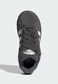 Mustat mokka-nilkkurit, joissa on kolme valkoista raitaa, teksturoitu nauhoitus ja pyöreä metallinen logo kielekkeessä.