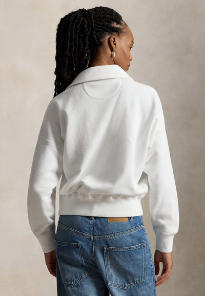 Γυναίκα με σκούρα πλεξούδα, που φοράει λευκό φούτερ με γιακά και μπλε τζιν, από πίσω, μπροστά σε απλό φόντο.