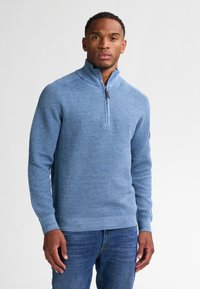 Lichtblauwe gebreide pullover trui met een kwart-rits kraag en ribbelstructuur, gecombineerd met denim jeans, met de armen ontspannen aan de zijkanten.