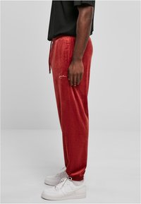 Red velour joggers featuring a drawstring waist, side pockets, and an embroidered logo. Paired with white sneakers.