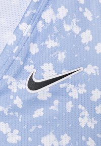 Zbliżenie na jasnoniebieską tkaninę z perforacjami w białe wzory przypominające chmury oraz czarne logo Nike w kształcie „swoosh”.